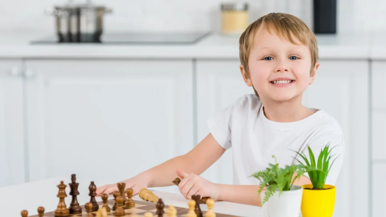 adorable smiling preschooler boy sitting at table, looking at camera and playing chess at home