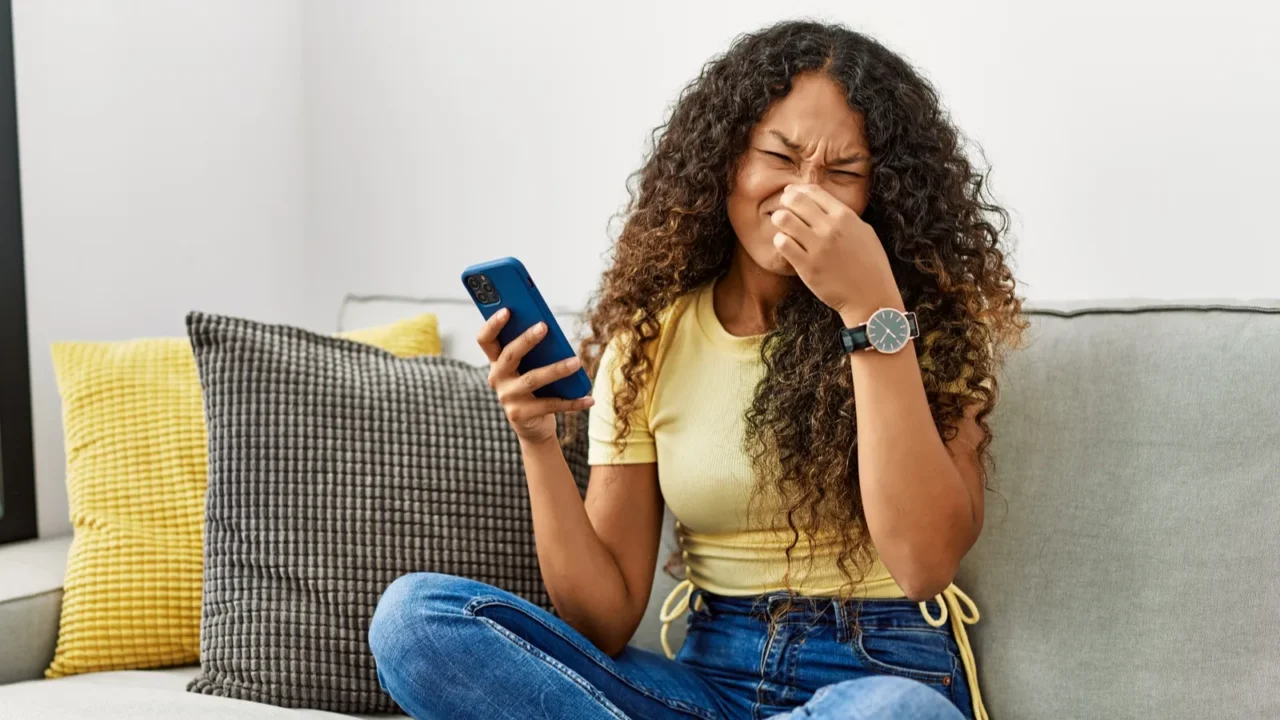 Young curly haired African woman sitting on the sofa in her living room while holding her nose with one hand because of the bad smell.