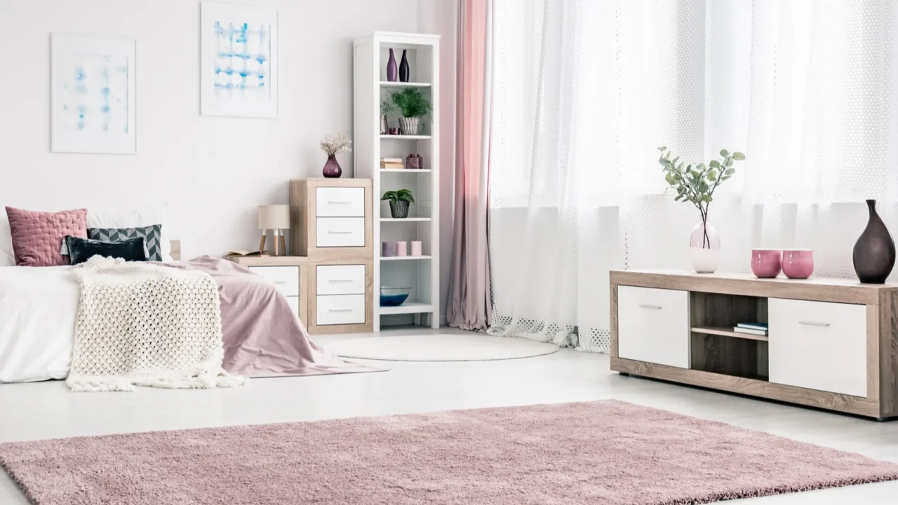 A pink bedroom interior with pink fluffy rug, bed with throw blanket and pillows, and shelf with decor accents.