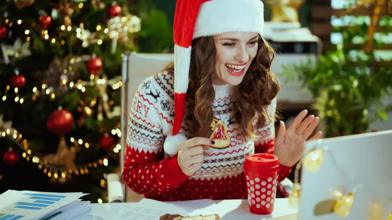 A woman dressed in Christmas clothes talking through a laptop in Christmas decorated home office.