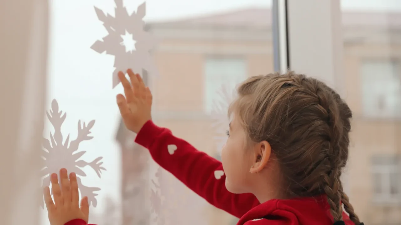 cute little girl decorating window with paper snowflake indoors