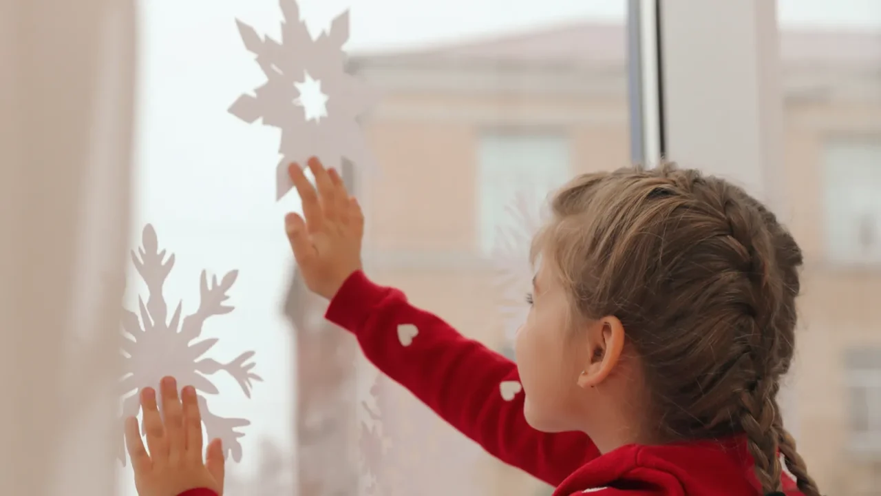 cute little girl decorating window with paper snowflake indoors