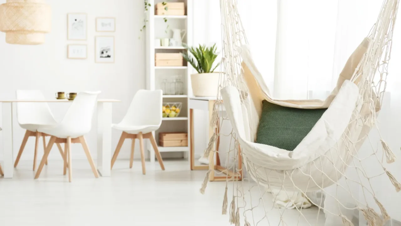 Macrame hammock swing in a white living room. Sofas, white chair, white shelf and plants. Organizers in shelf. Hanging rattan wicker light and frames hanging on the wall. A well-lit room. White sheer curtains.