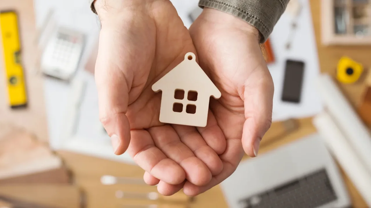 Close-up of hands cupping a small wooden house model on a cluttered work surface