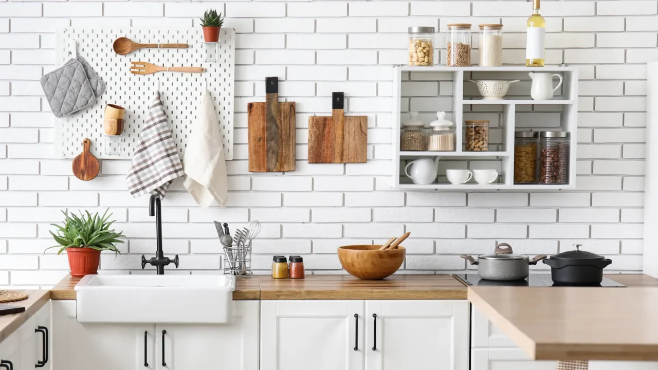 Kitchen counters with utensils and wooden board hanging on the wall. Shelf unit on white bricks.