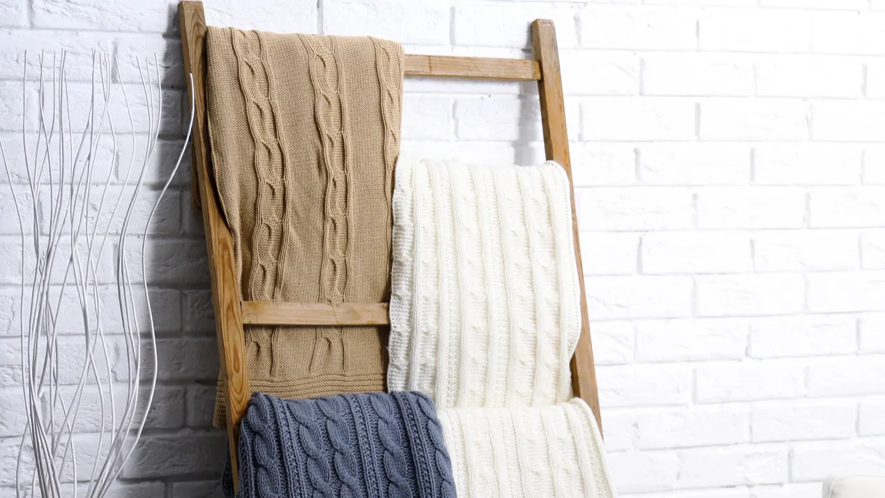 A wooden ladder with throw blankets on it against a white brick wall.