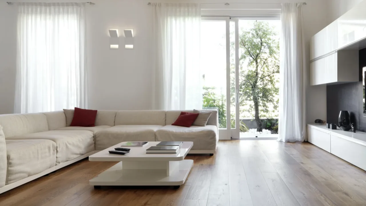 Modern living room with wood floor, white furniture, red cushions, and large windows.