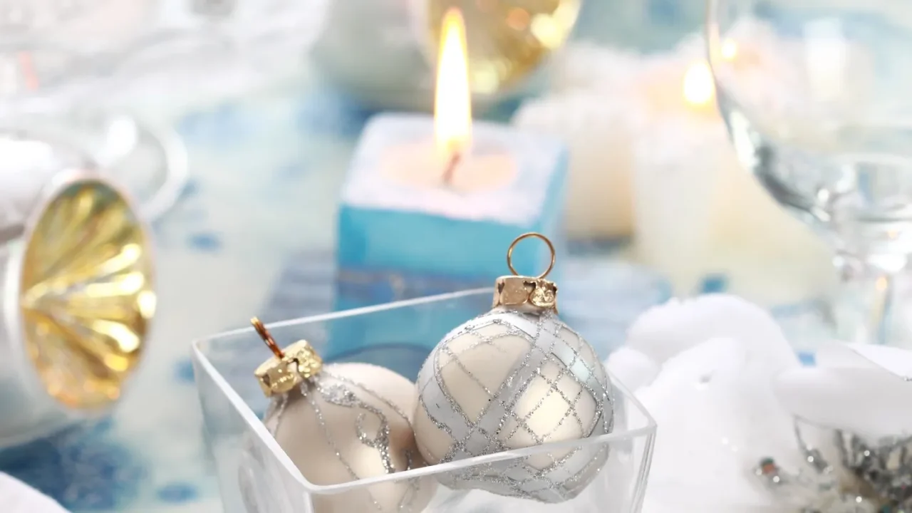 Blue and gold candles, baubles and decor for Christmas. A lit candle and glasses in the background. White, blue, silver, and light gold decor.