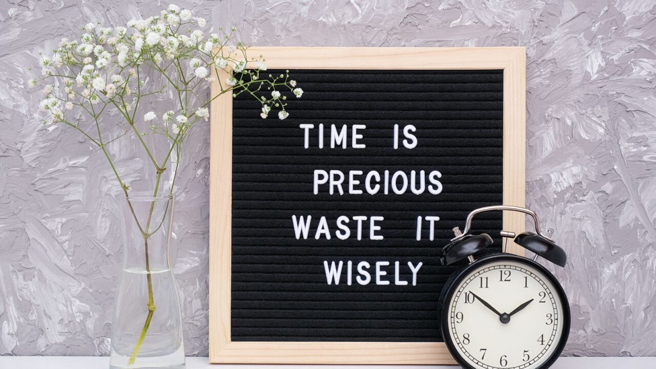 Time is precious waste it wisely. Motivational quote on letterboard, black alarm clock, flower in vase on table against stone wall. Concept quote of the day.