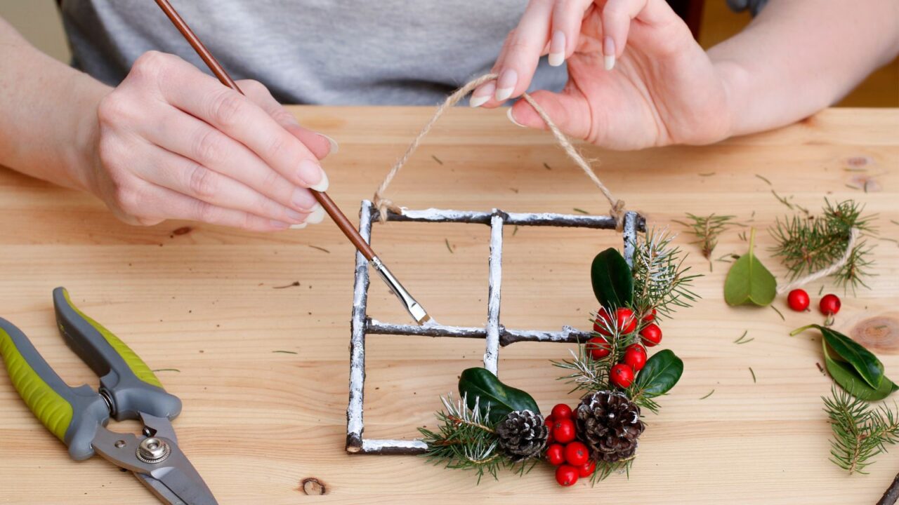 Florist at work: woman shows how to make miniature wooden window frame with twigs decorated with holly (ilex), fir and cones. Traditional Christmas decoration.