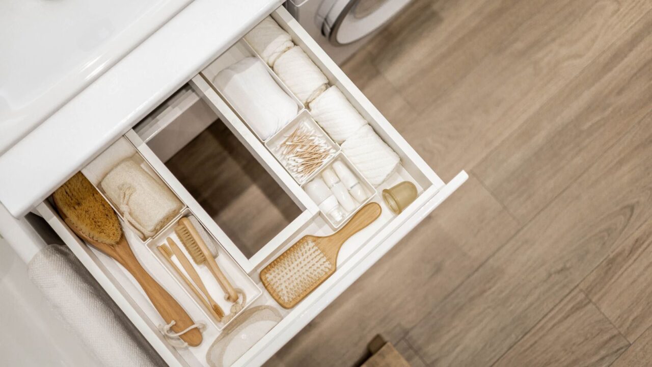 Bathroom under sink organizer drawers with neatly placed bath amenities and toiletries. Table top basin sink with dispenser of liquid soap, toothbrush in cup and group of rolled up white towels.