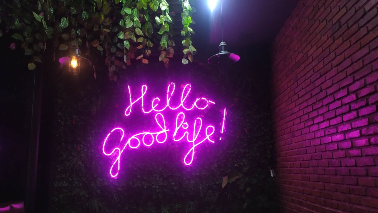 "Hello Good Life" led neon sign on a leaf-wall. Hanging lights can be seen. Bricked wall on the right.