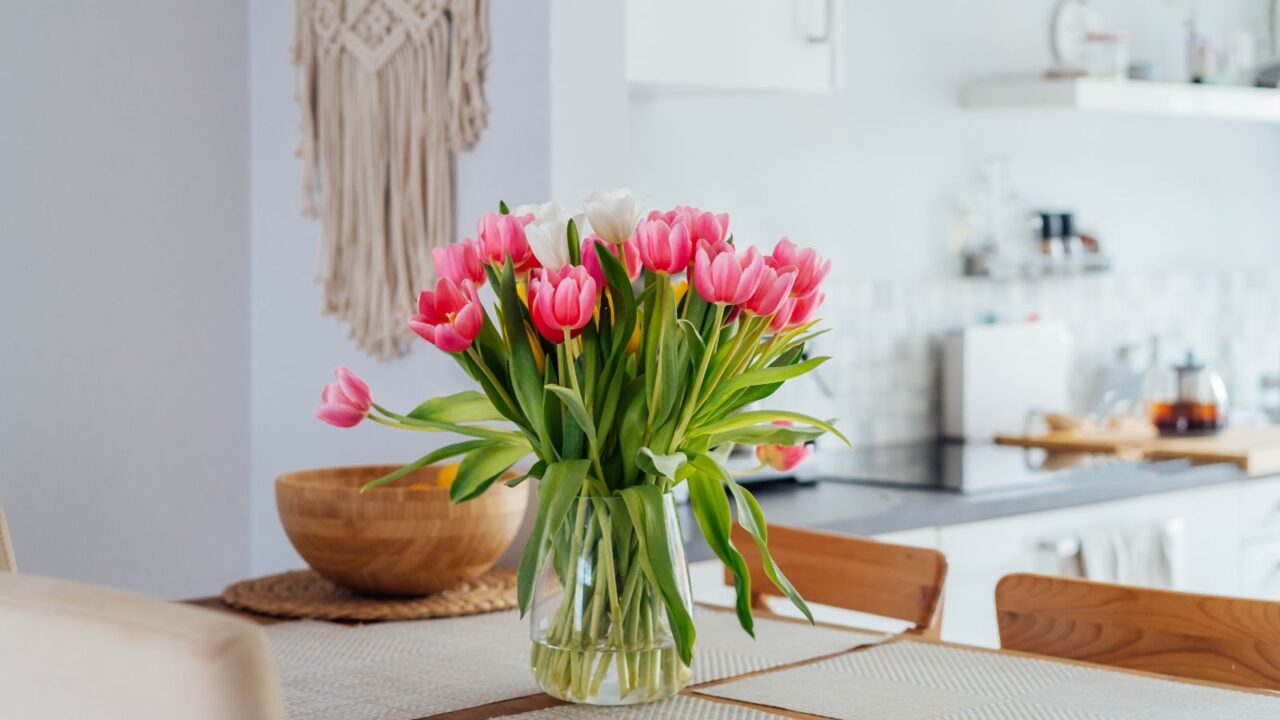 Stylish and modern boho, Scandi interior of open space. White kitchen with pink tulip flowers in vase on the wooden kitchen counter table, macrame on the wall. Cozy design home decor. Selective focus.