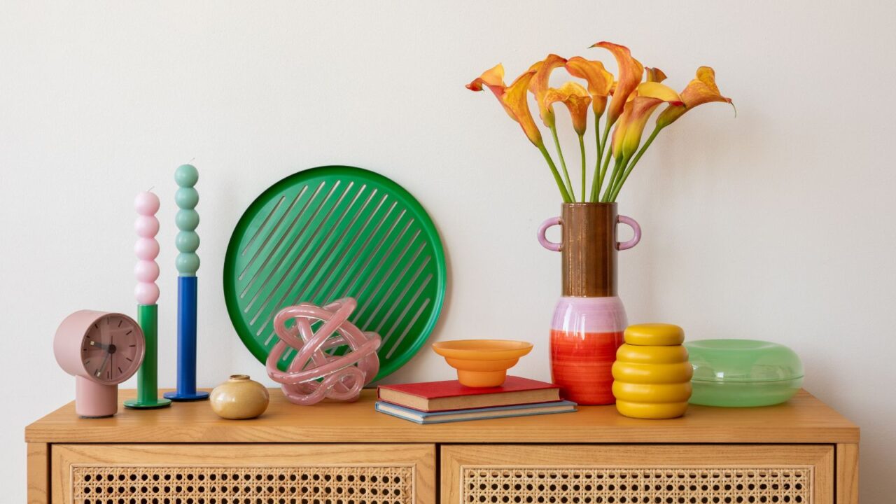 Modern tabletop decor with orange calla lilies in a ceramic vase, colorful candles, and abstract decor pieces on a wooden cabinet, creating a vibrant and stylish interior scene.