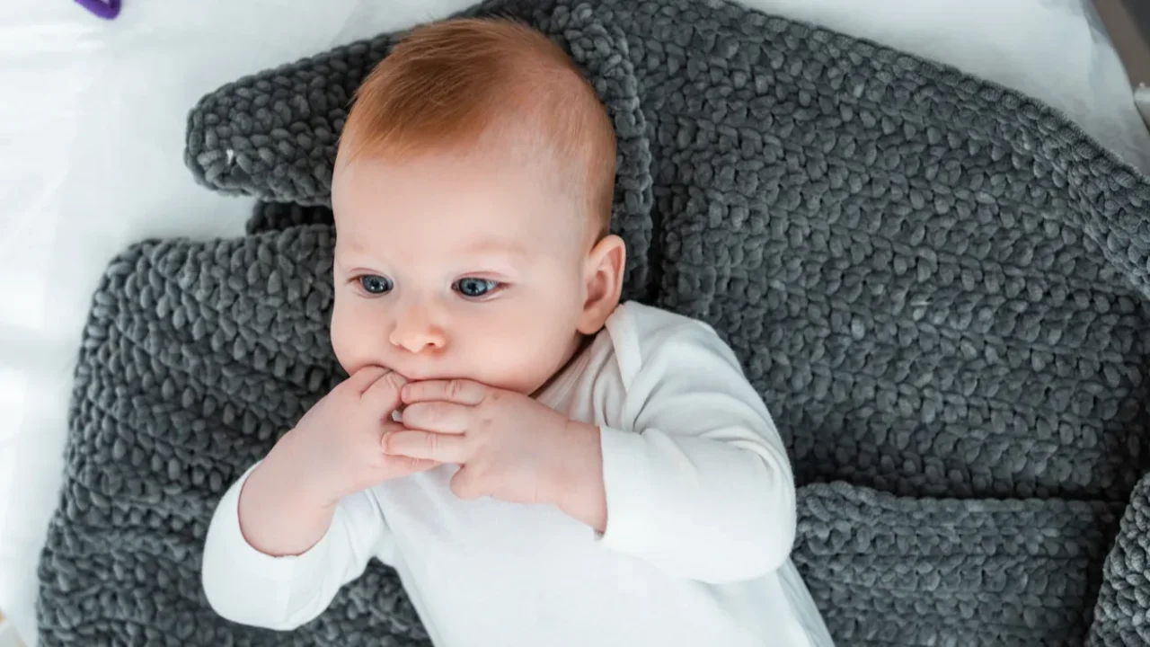 top view of adorable baby boy lying in baby cot on blanket with hands near mouth