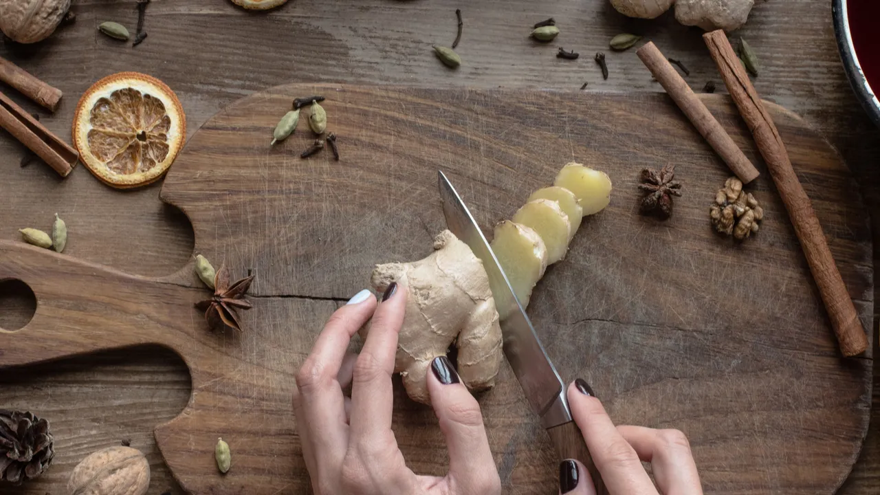 Woman cutting ginger on a wooden board. Cinnamon, cloves, star anises, and dried citrus are also placed.