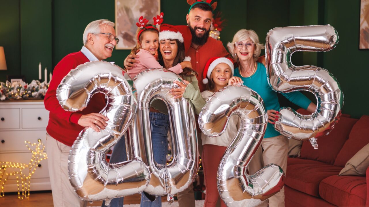 A family celebrates New Year's 2025 with giant silver balloons shaped like the numbers 2, 0, 2, and 5, all smiling and wearing festive accessories.