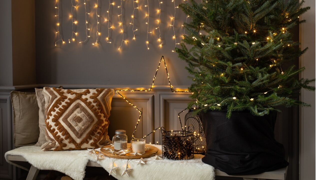 A cozy holiday scene with a decorated Christmas tree, string lights, a star-shaped light, and a tray with candles and a jar on a bench with pillows and a fur throw.