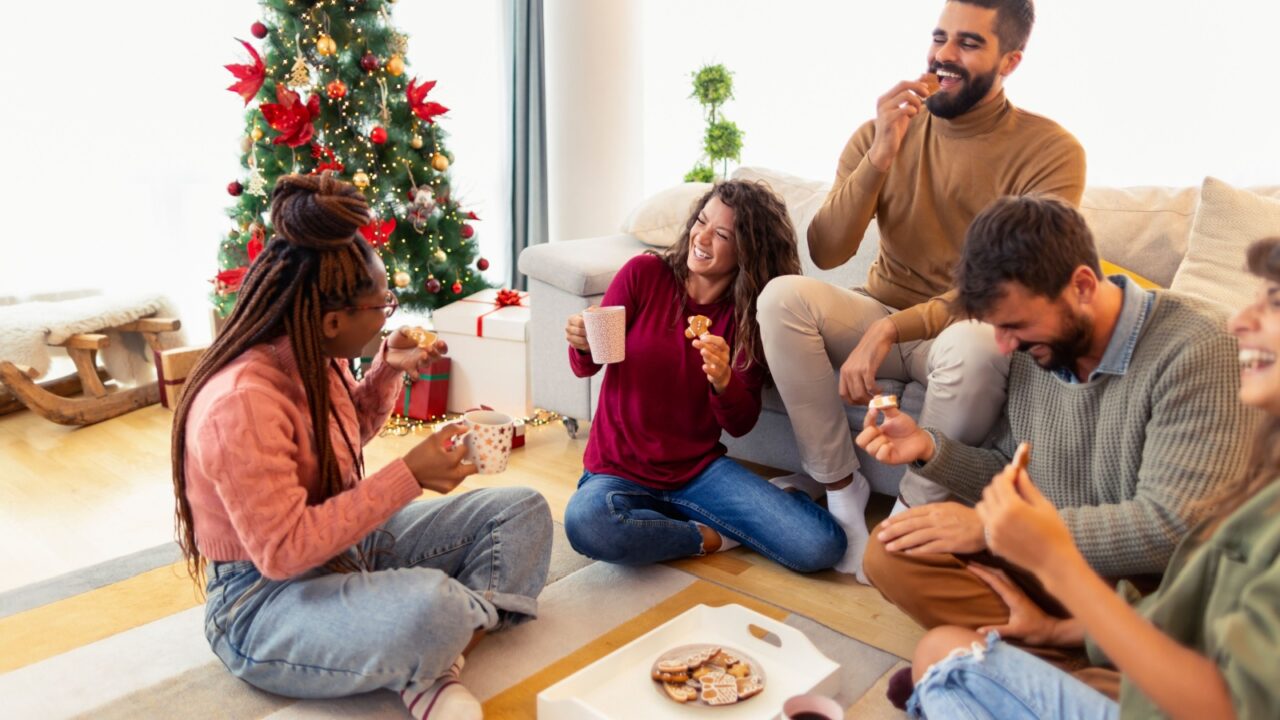 A cozy group of friends enjoying snacks and drinks around a coffee table, with a decorated Christmas tree in the background.