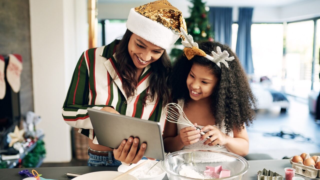 Mother and daughter in festive attire baking together while looking at a tablet.