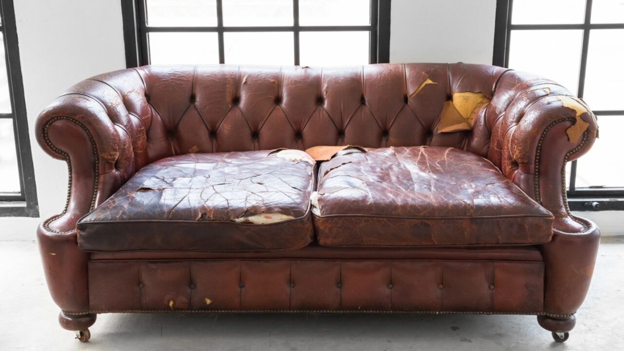 A worn-out, brown leather sofa with visible tears and cracks in the upholstery, placed in front of large windows.