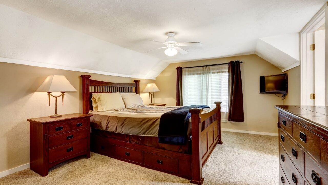 A cozy low-ceiling bedroom with a wooden bed, nightstands, lamps, a dresser, and a wall-mounted TV.