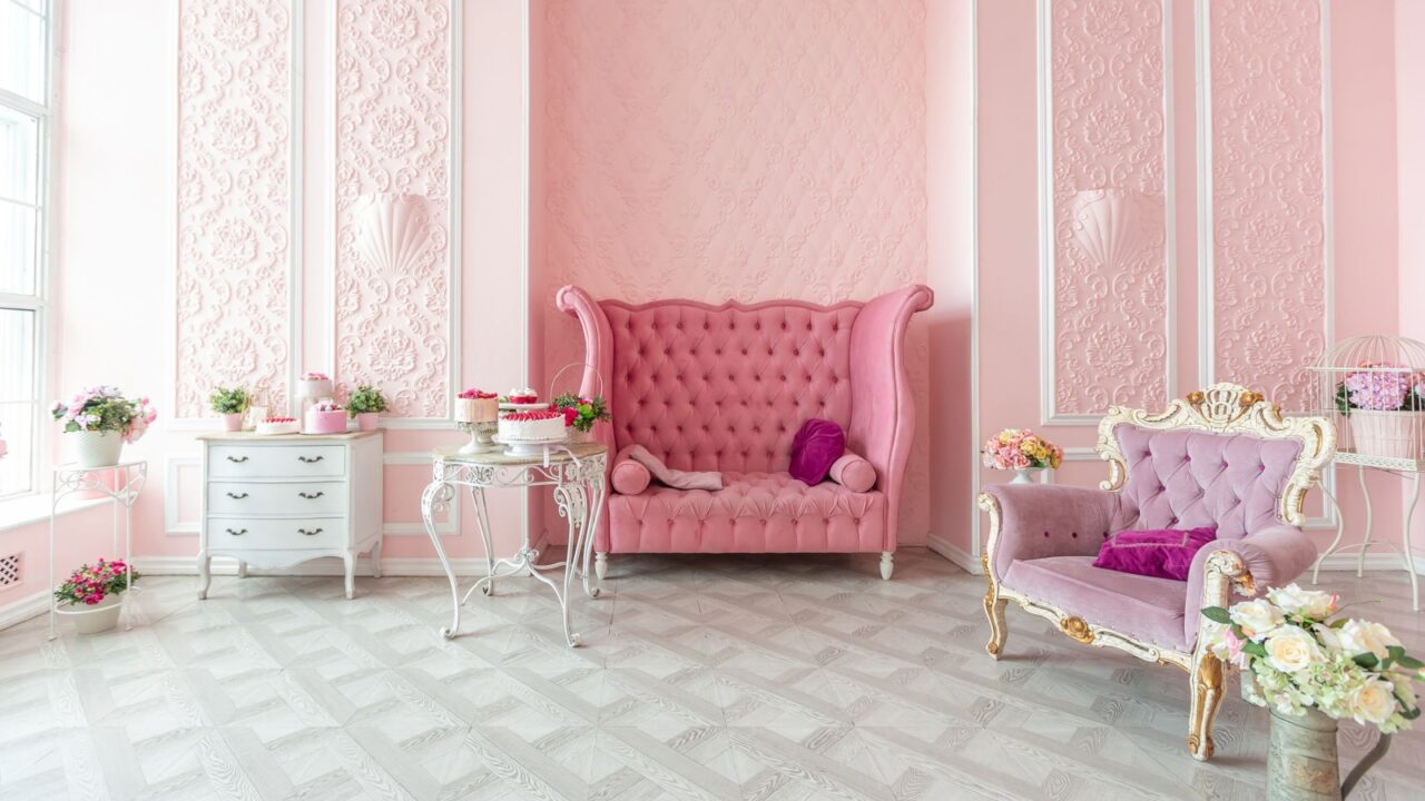 A luxurious pink-themed room with ornate furniture, including a tufted high-back sofa, a smaller armchair, and various floral decorations.