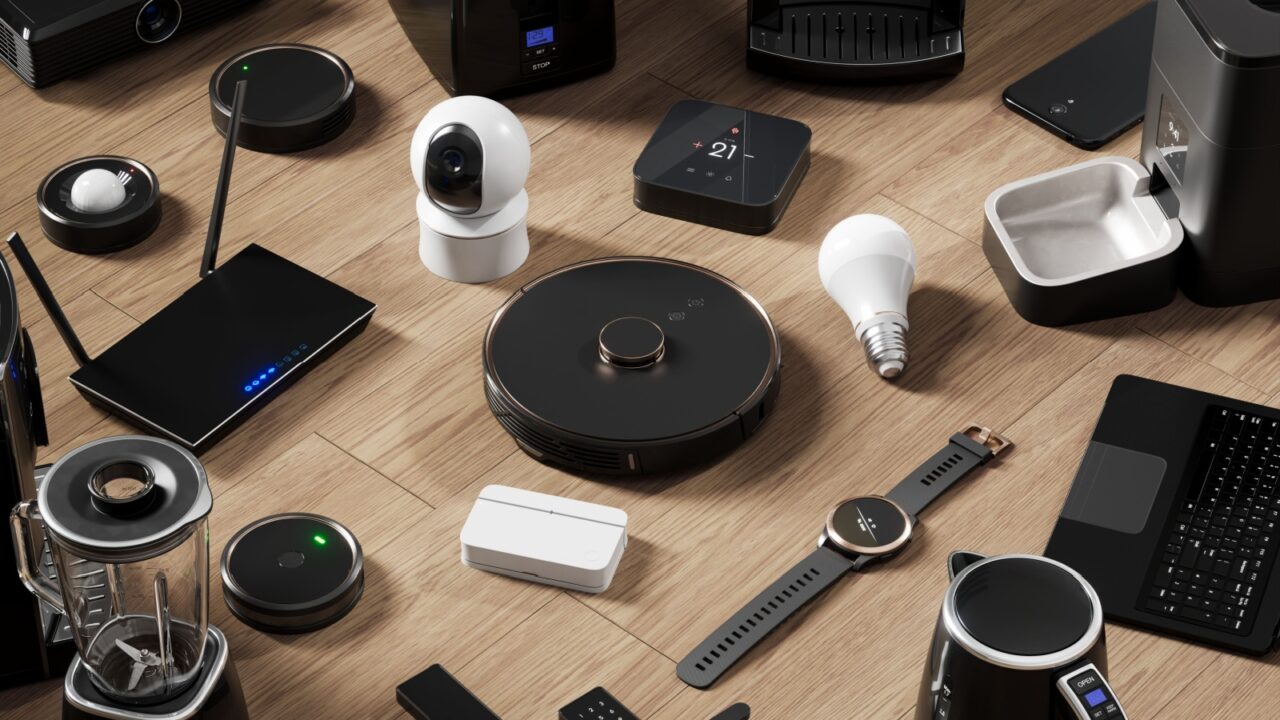 A collection of various smart home devices arranged on a wooden floor, including a robot vacuum, smart light bulb, smart thermostat, security camera, smartwatch, and other electronic gadgets.