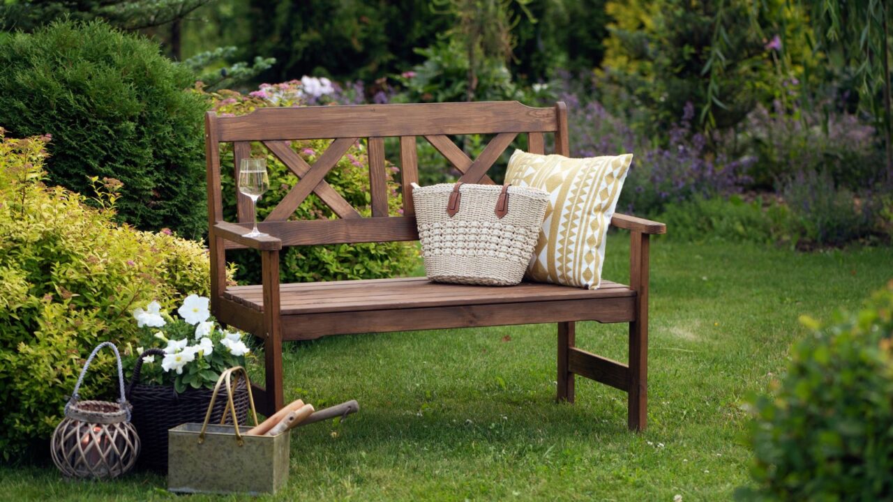 A wooden bench in a garden with a basket, cushion, and potted flowers next to it.