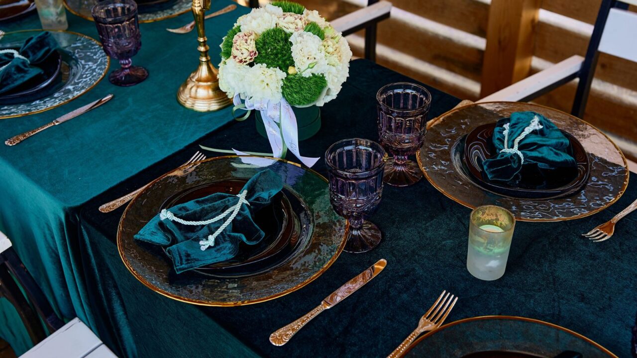 A dining table setup with blue velvet tablecloth, velvet napkins on plates, glasses, and a flower vase.