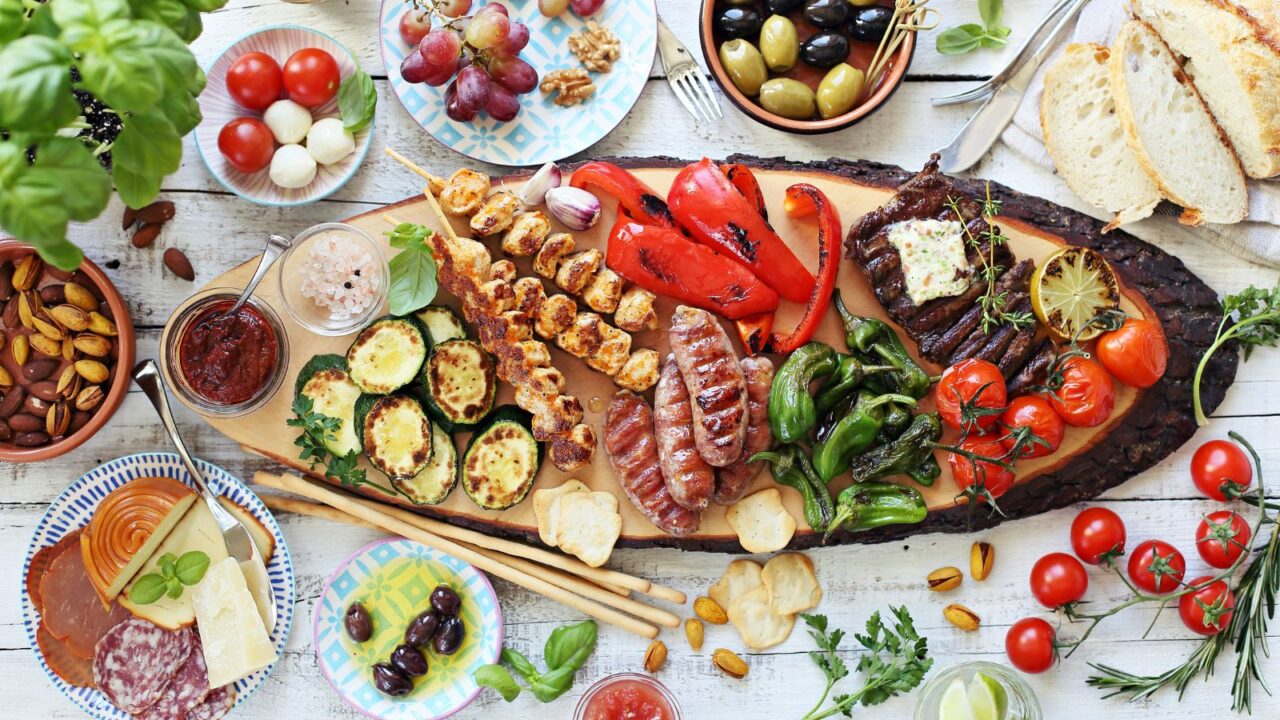 A wooden platter with grilled meat, chicken skewers and sausage with roasted vegetables and appetizers on a wooden table.