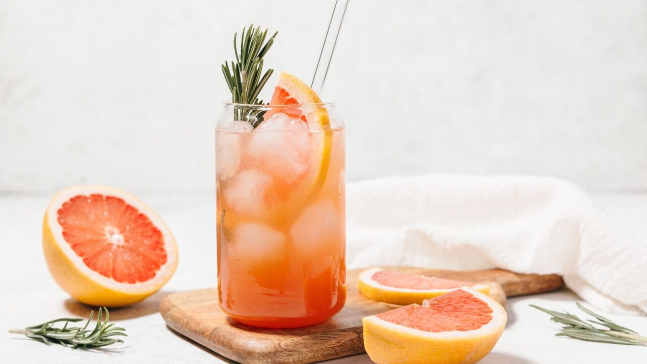 A grapefruit cocktail with rosemary garnish in a glass, served on a wooden board.