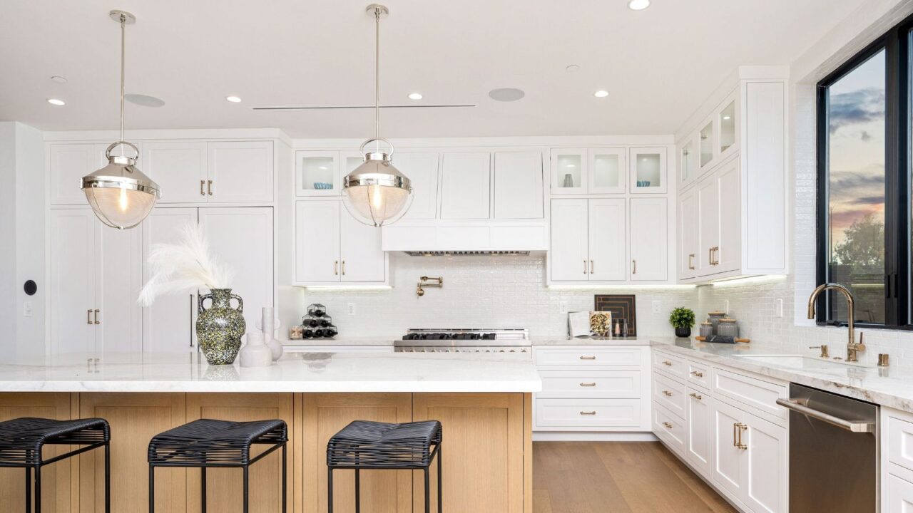 A kitchen interior with white cabinets, granite countertop, window, bar stools, and pendant lights hanging from the ceiling.