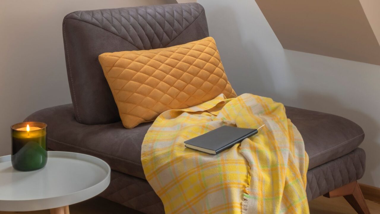 A cozy reading nook with throw blanket and pillow on a couch and a white table with a candle on it.