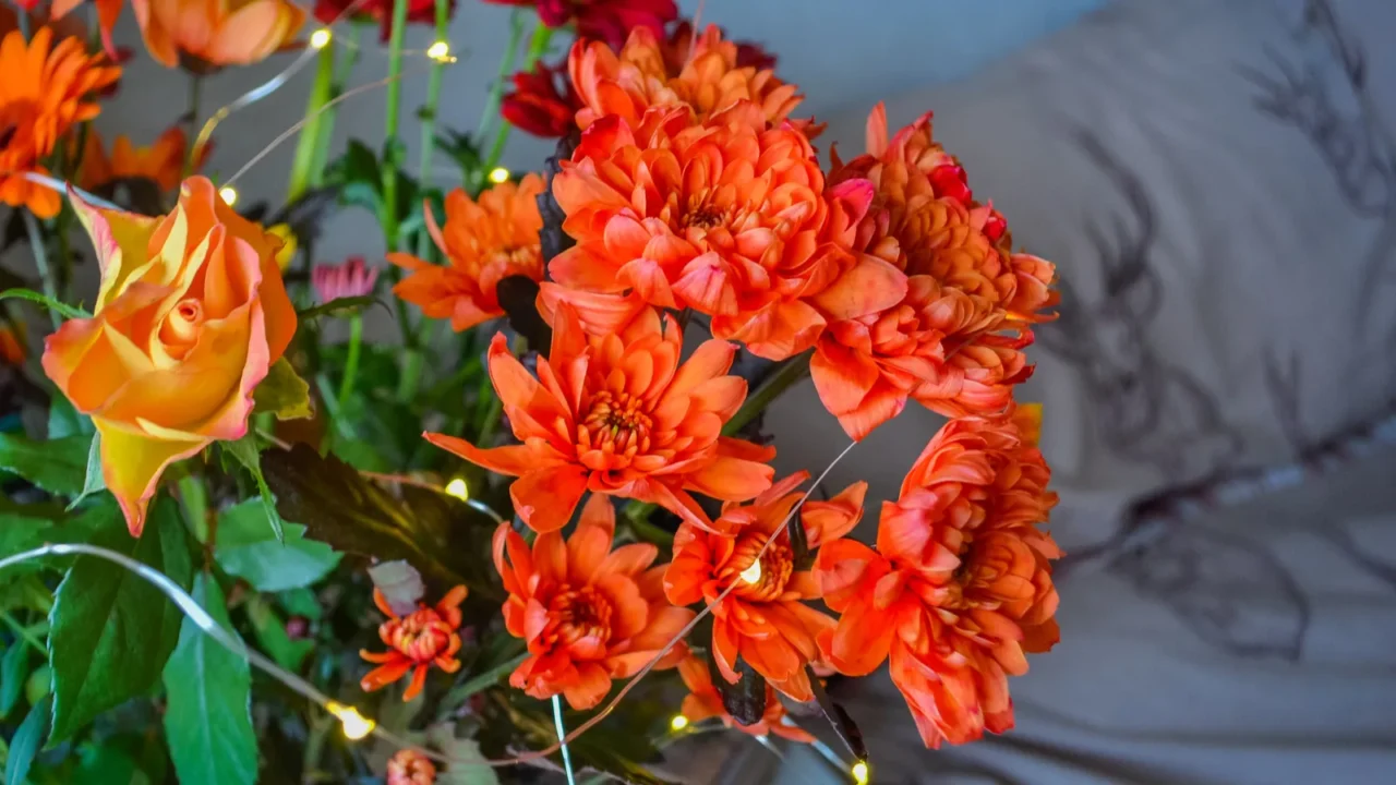 A bouquet of fresh orange and yellow flowers including carnations and roses at home with fairy lights around it.