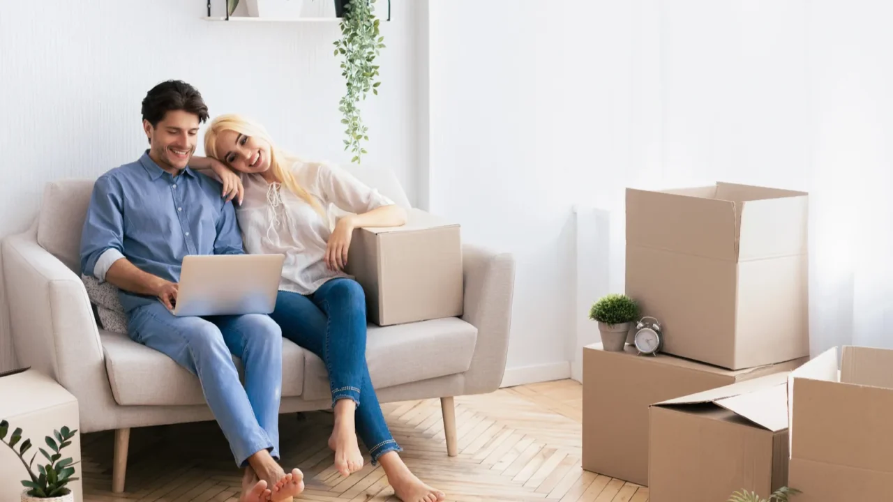 A couple sits on a sofa with a laptop, surrounded by cardboard boxes in a bright, cozy room.