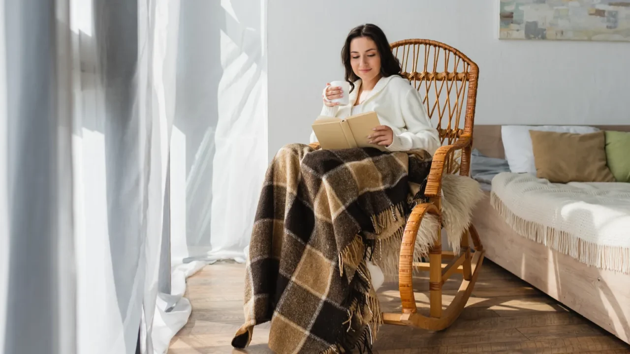 A woman reading a book in a cozy nook near the window in a rocking chair with a throw blanket and mug.