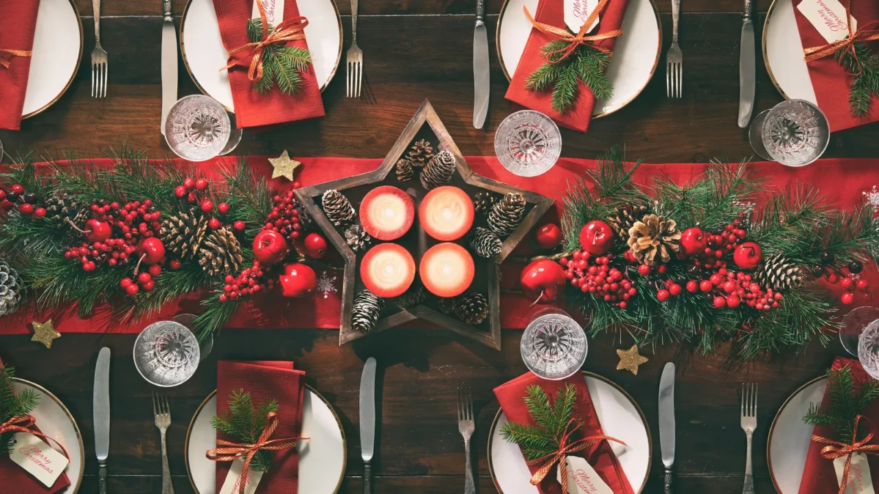 A wooden table with red table runner, plates with napkins, glassware, and candles.