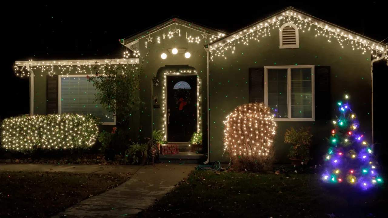 Exterior of house decorated with bright Christmas lights, glowing bushes, and a colorful tree.