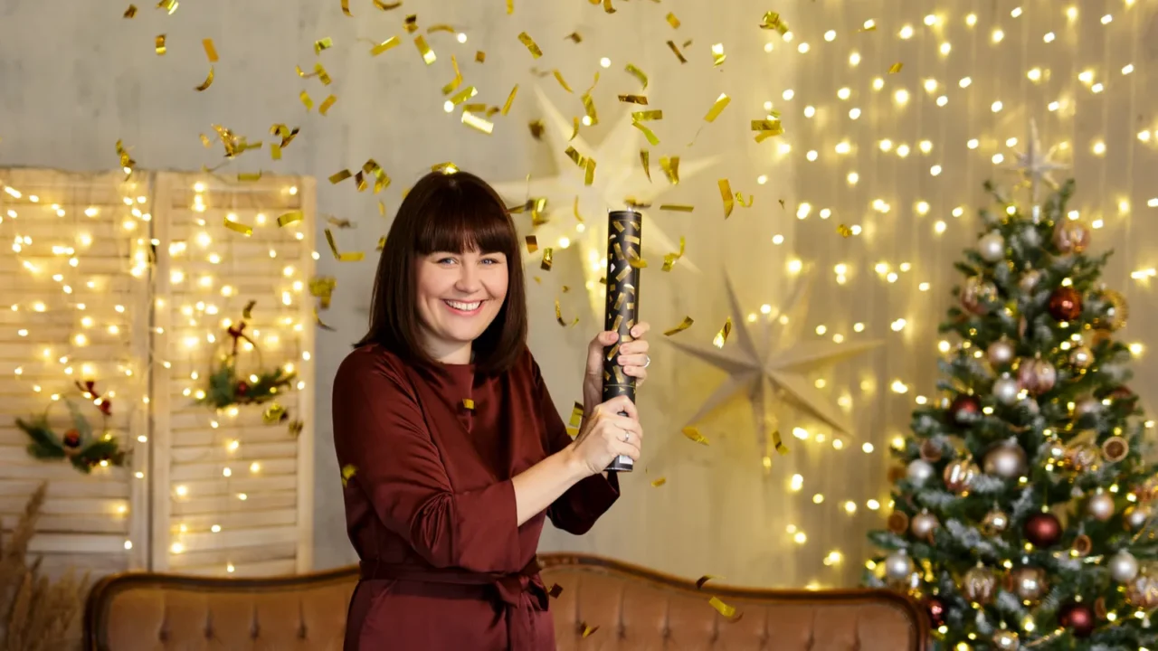 A happy young woman holding a confetti popper in a decorated living room with a Christmas tree and garland lights.