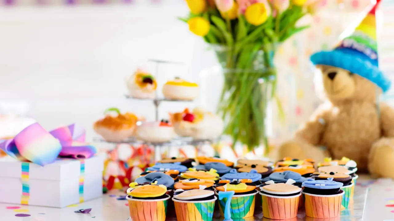 Cupcakes for kids. Rainbow-themed birthday party with confetti, gifts, ribbons, and rainbow hats. A teddy wearing a rainbow hat in the background.