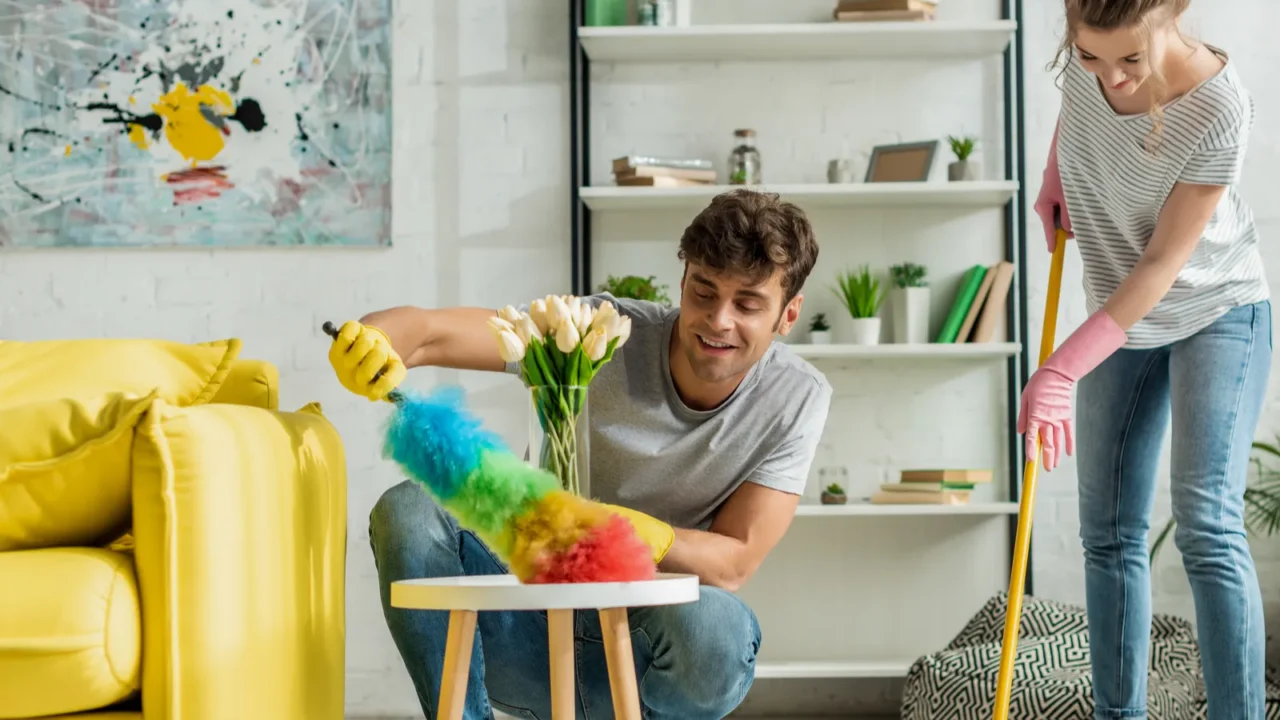 Two people cleaning a room; one uses a colorful duster while the other mops.