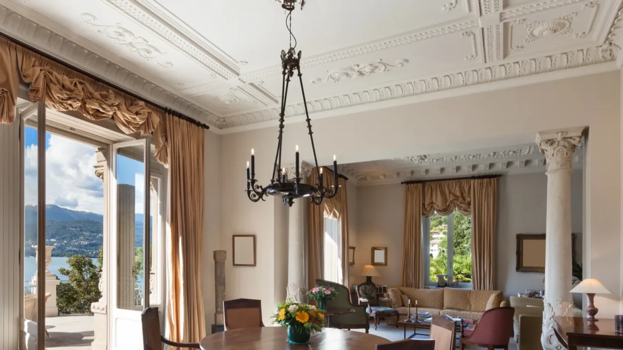 Luxury living room interior with white textured plaster ceiling. A black chandelier, a sofa set, a dining table, and a big door and windows with curtains on it are all a part of the room.