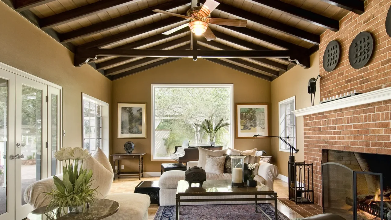 A big living room with exposed beams, decor, sofa set, and large windows. Well-lit space. Frames hanging on the wall with planters and some antique decor. A firepit and 3 black wall hangings.