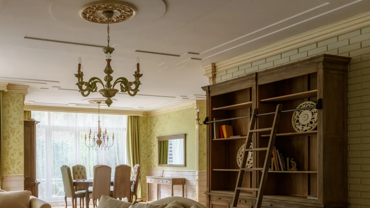 Living room interior with chandeliers, a dining table in a soft off white tone, a bricked wall on the left with a weathered wood bookshelf and a ladder resting on it. A light green wallpaper with a mirror and a thin console table at the back.