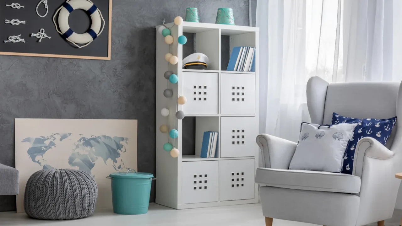 Living room with nautical decor. Shelves with open and closed drawers and a small blue bucket on the floor for storage and a grey pouf for seating. A grey sofa with cushions.