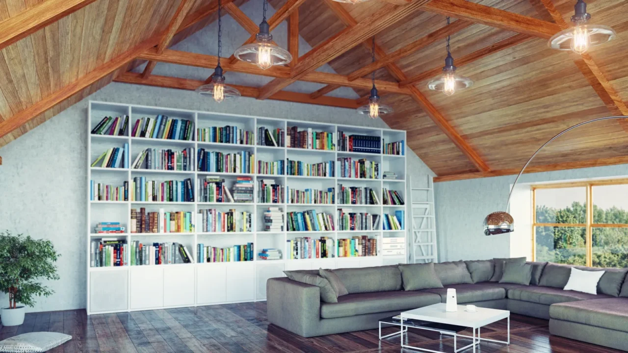 Modern attic with a wooden beams ceilings. A big white bookshelf on the wall and a window to the right. A floor lamp, gray sofa, and a white table on dark brown wooden flooring. Lights are hanging from the ceiling and an indoor plant is placed on the left.