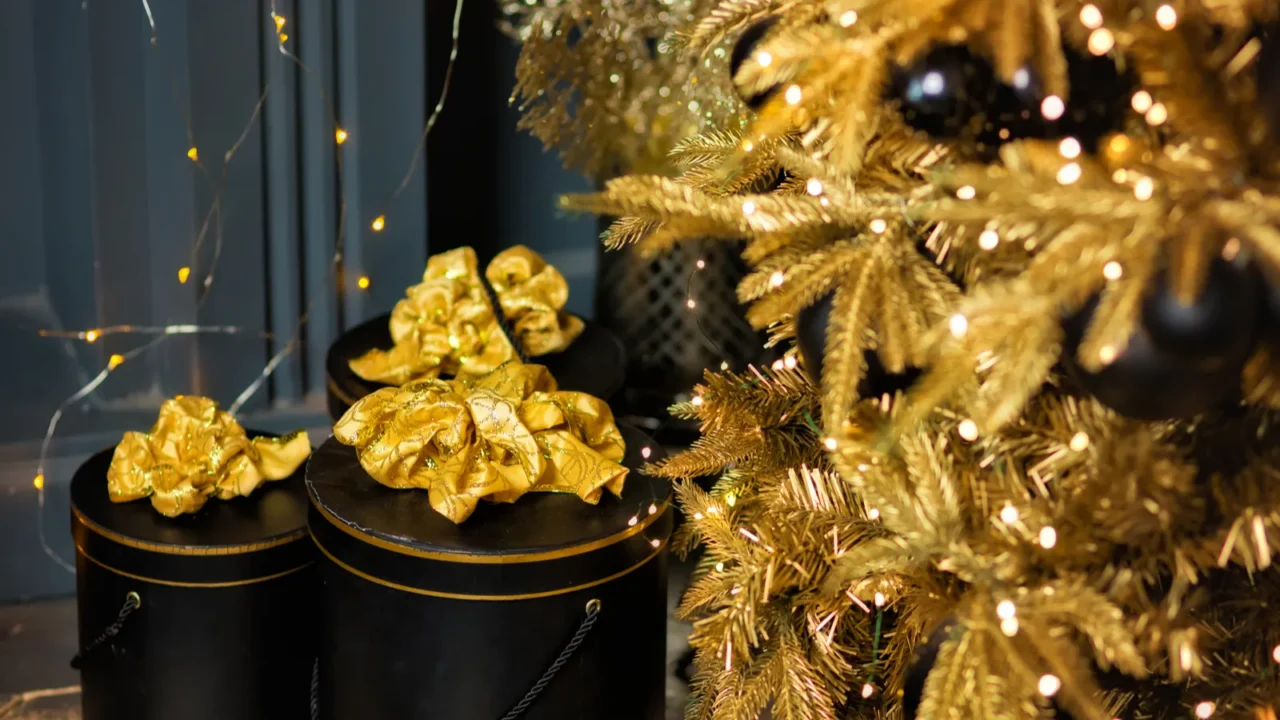 Three black gift boxes with gold ribbons stacked near a beautifully decorated gold Christmas tree, glowing with lights.