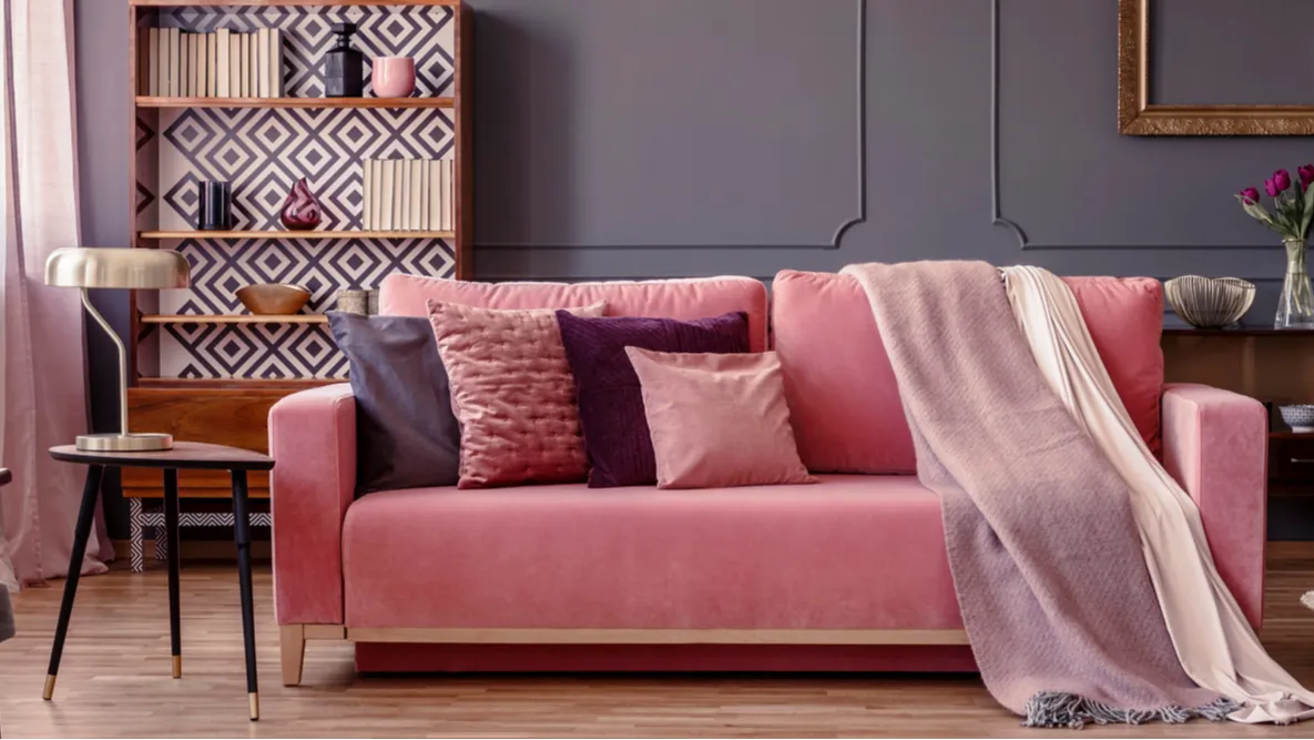 Powder pink velvet couch with decorative pillows and two throw blankets. A coffee table and shelf.