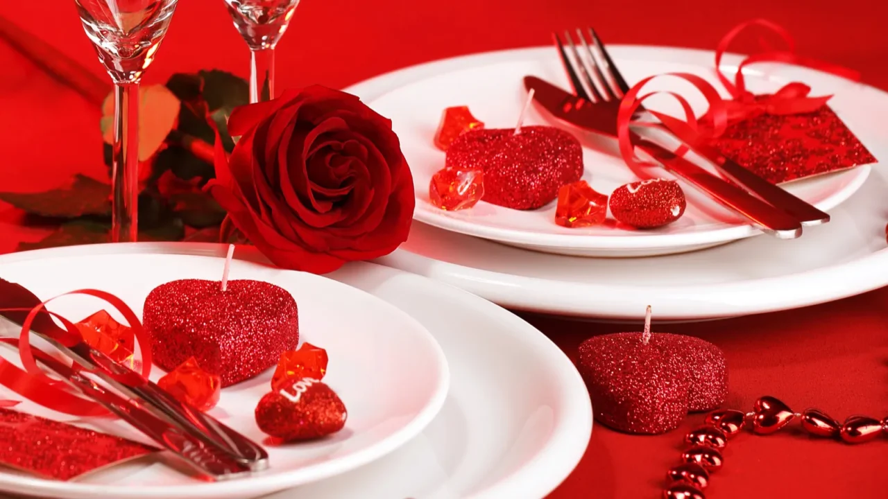 Romantic dinner table with red tablecloth. White plates, a rose, wine glasses, and candles for decor.